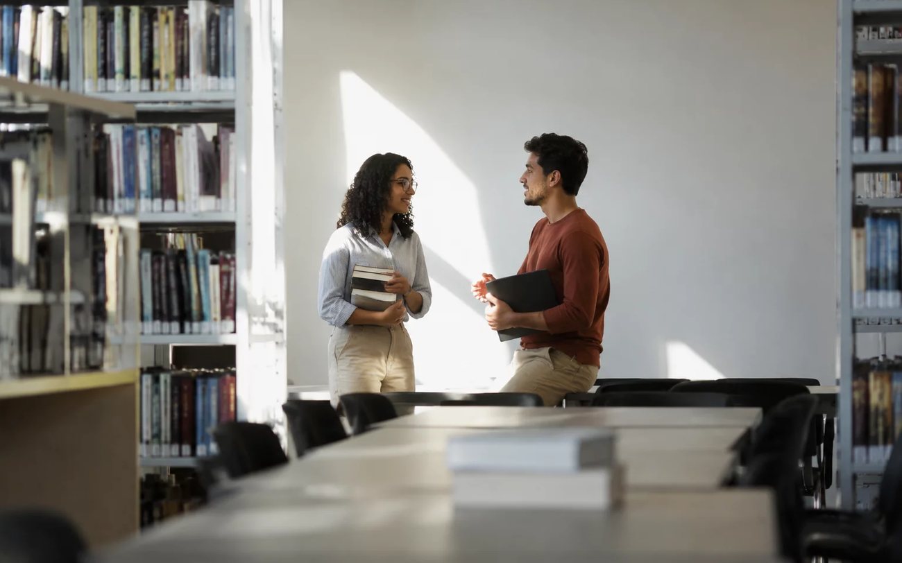 Estudantes conversam em pé no corredor entre as estantes de uma biblioteca